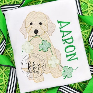 May include: Embroidered design of a light brown dog holding a string of green shamrocks. The dog is sitting and the name "AARON" is embroidered in green letters on the right side of the design. The design is on a white fabric.