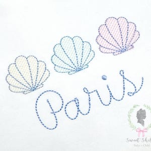 May include: Embroidered design featuring three seashell motifs in cream, light blue, and pink, with the word "Paris" in a cursive font. The design is on a white background. A logo with a silhouette and text is in the bottom right corner.