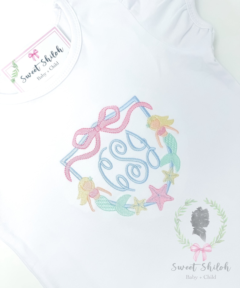 May include: A white shirt with a light blue, pink, and green embroidered monogram surrounded by mermaids and starfish. The monogram is in a square frame with a pink bow at the top. The text 'Sweet Shiloh Baby + Child' is on a pink tag in the top left corner of the shirt. The text 'Sweet Shiloh Baby + Child' is also on the bottom right corner of the shirt.