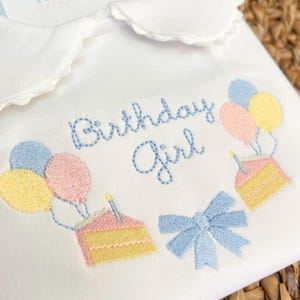 May include: White embroidered baby onesie with blue and pink balloons, a cake with candles, and a blue bow. The onesie says "Birthday Girl" in blue embroidery.