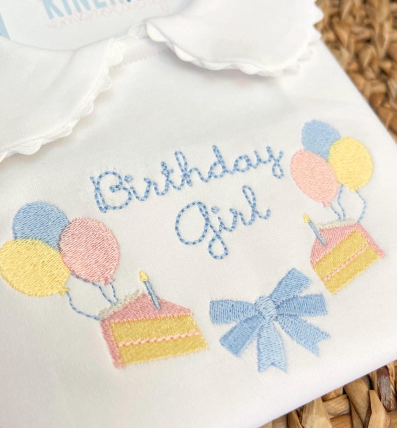 Op de afbeelding: Witte geborduurde baby romper met blauwe en roze ballonnen, een taart met kaarsen en een blauwe strik. De romper zegt "Birthday Girl" in blauwe borduurwerk.