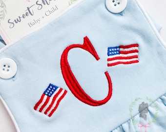 May include: Pale blue baby romper with white edging, embroidered with a large red letter "C" and two small American flags. The romper has white buttons and the text "Sweet Shiloh Baby + Child" on a card.