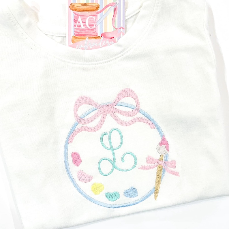 May include: Pastel-colored embroidered sweatshirt featuring a circular design resembling a painter's palette. The palette includes a light-green initial 'L', a paintbrush, and a pink bow.  The artwork is stitched onto a white sweatshirt. A small tag shows the embroidery brand logo.