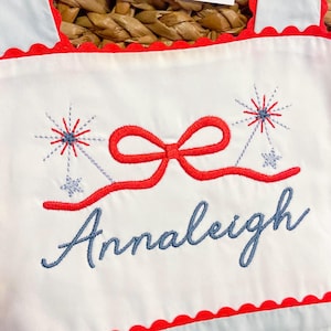 May include: White fabric with red scalloped trim and straps. Features a red embroidered bow and fireworks design, with the name "Annaleigh" in blue script. The design is centered on the fabric.