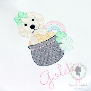 Könnte beinhalten: Gesticktes Design mit einem cremefarbenen Welpen mit grüner Schleife, der aus einem schwarzen Topf voller Goldmünzen auftaucht. Ein Regenbogen und ein Kleeblatt sind ebenfalls vorhanden. Der Name "Goldi" ist in rosa Schrift gestickt.