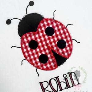 May include: A white background features a ladybug design with a red and white gingham pattern on its wings and black spots. The ladybug has black antennae and legs. The name "ROBIN" is embroidered below the ladybug, with the text "Sweet Shiloh Baby + Child".