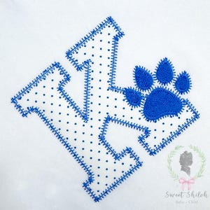 May include: White fabric with a blue stitched letter "L" and a blue paw print. The letter is white with blue polka dots and a blue stitched outline. The paw print is solid blue with a blue stitched outline. The text "Sweet Shiloh Baby Child" is in the bottom right corner.