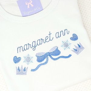 May include: White baby onesie with blue embroidery of a bow, snowflakes, crowns, and hearts. The onesie has the name "margaret ann" embroidered on it.