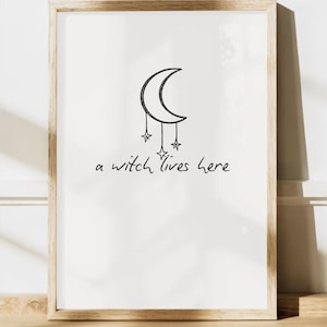 May include: A framed print featuring a crescent moon and three hanging stars, with the handwritten text "a witch lives here" in black. The artwork is set against a white background and framed in light brown wood.