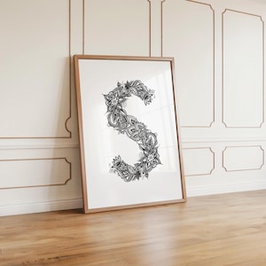 Könnte beinhalten: Gerahmter Druck des Buchstabens "S" mit einem dekorativen schwarz-weißen Blumenmuster. Das Kunstwerk befindet sich in einem hellbraunen Rahmen und lehnt an einer weißen Wand. Das komplizierte Design ist ein auffälliges Merkmal des Drucks.