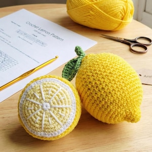 Op de afbeelding: Twee gehaakte citroenen, één heel en één doorgesneden, met groene bladaccenten. Een bol geel garen, een haaknaald, een schaar en een patroon met de titel "Crochet Lemon Pattern" zijn ook zichtbaar. De citroenen zijn geel met witte en lichtgele details.