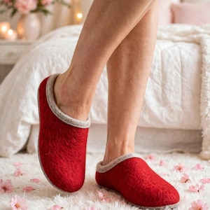 May include: A pair of red felted wool slippers featuring a textured exterior and a light gray trim along the collar and sole. The slippers have a rounded toe design and a slip-on style, providing full coverage for the foot while maintaining a soft, dense appearance.
