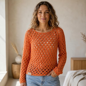 Op de afbeelding: Een oranje gehaakte top met lange mouwen en een ronde halslijn. Het open weefsel onthult de huid eronder. De top wordt gecombineerd met een blauwe spijkerbroek. Een klein label is zichtbaar aan de linkerkant van de top.