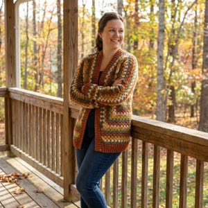 Puede incluir: Un cárdigan de ganchillo multicolor en tonos marrón, naranja, verde y amarillo. El suéter abierto tiene un patrón texturizado y lo lleva una persona en un porche de madera. El fondo presenta follaje otoñal.