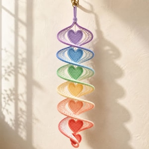 May include: A vertical hanging spiral decoration made of crocheted yarn featuring a gradient of colors including purple, blue, green, yellow, orange, and red. Each section of the spiral contains a solid crocheted heart in a matching color.