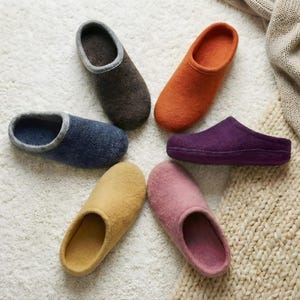 Peut inclure: Six pantoufles colorées, disposées en cercle sur une surface blanche texturée. Les pantoufles sont de différentes couleurs : marron, bleu, orange, violet, jaune et rose. Elles semblent être faites d'une matière douce, évoquant le confort.