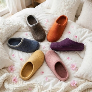 Può includere: Sei pantofole in lana infeltrita disposte a cerchio su una superficie bianca testurizzata. Le pantofole sono disponibili in colori solidi tra cui marrone scuro, arancione, blu scuro, viola, giallo e rosa polvere, con un semplice design a infilare.