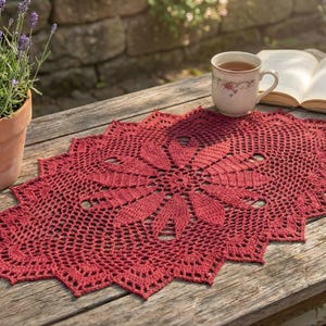 May include: A handmade, red crochet doily with a floral design, resting on a weathered wooden table. A cup of tea, a book, and a potted lavender plant are also on the table, creating a cozy outdoor scene.