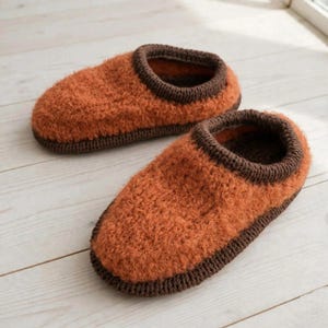 May include: A pair of slippers featuring a fuzzy, textured orange upper and a dark brown knitted trim around the opening and the base. The slippers have a rounded toe design and a soft, plush appearance, showcasing a two-tone colour scheme.