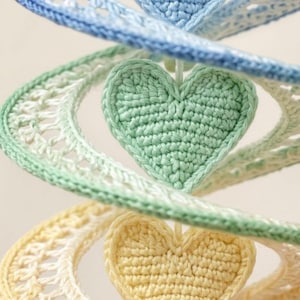 May include: A vertical spiral mobile featuring three crocheted hearts in pastel blue, mint green, and pale yellow. Each heart is attached to a crocheted ribbon with an intricate openwork lace pattern that transitions in color, creating a soft gradient effect.