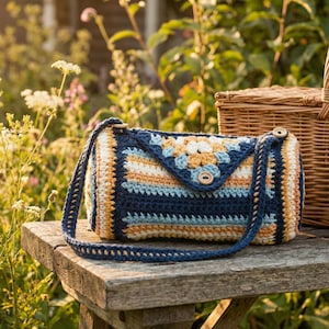 May include: A handmade crochet handbag in a rectangular shape with a flap closure. The bag features a color block design with navy blue, yellow, and white stripes. The strap and button closure are navy blue. The bag is sitting on a wooden surface.
