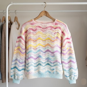 Op de afbeelding: Een gehaakte trui met lange mouwen en een herhalend golfpatroon in een pastelkleurig regenboogverloop van roze, geel, oranje, paars en blauw op een witte basis. Het kledingstuk heeft een ronde halslijn en geribbelde boorden aan de mouwen.