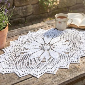 Pode incluir: Uma toalha de mesa branca de crochê com um design floral, em uma mesa de madeira desgastada. Uma xícara de chá e um livro aberto também estão presentes, junto com uma planta de lavanda em vaso. O padrão intrincado da toalha de mesa é visível.