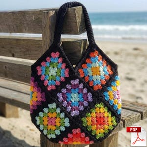 May include: A black crocheted tote bag with a colorful granny square design. The bag features a sturdy, braided handle and is displayed outdoors on a wooden bench near the beach. The bag's squares showcase a vibrant mix of colors.