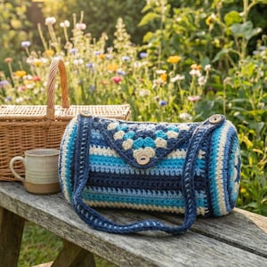 May include: A handmade crochet bag in shades of blue, cream, and navy, with a flap closure and wooden button detail. The bag has a long strap and is set on a wooden bench with a picnic basket and mug in the background.