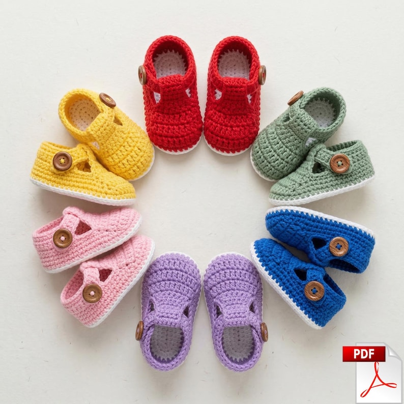 May include: A selection of colourful, handmade crochet baby shoes arranged in a circle. The shoes are in shades of yellow, red, green, pink, purple, and blue. Each shoe has a button fastening and a white sole. The image displays a variety of baby footwear.