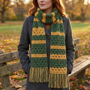 Puede incluir: Una bufanda de ganchillo verde y dorada con flecos. La bufanda presenta bandas alternas verdes y doradas, con un diseño calado texturizado. La bufanda se lleva sobre un abrigo gris, lo que sugiere un accesorio de otoño o invierno.
