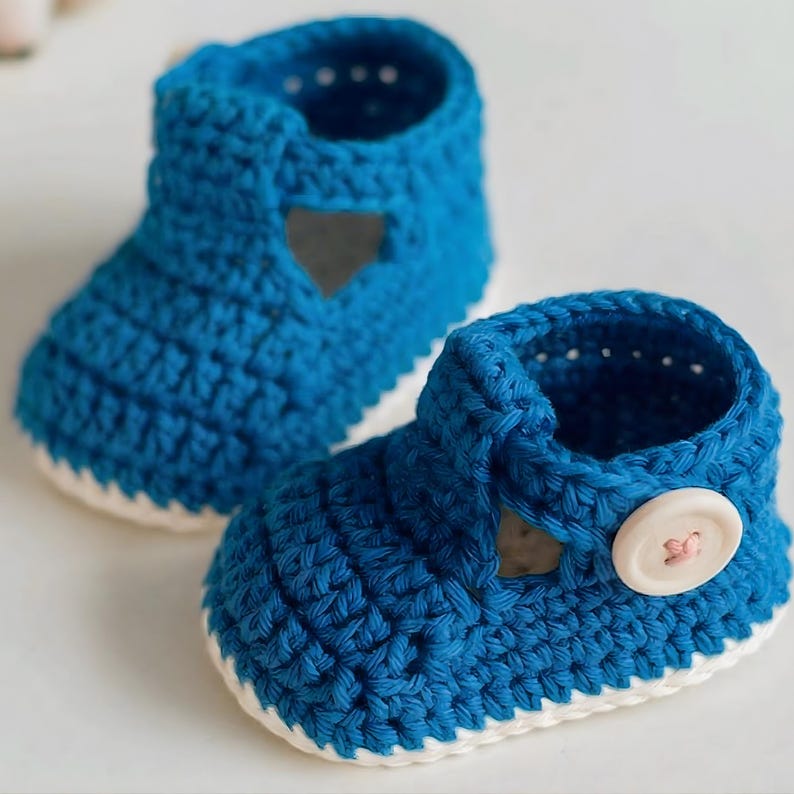 May include: A pair of blue crochet baby booties. The shoes have a white sole and a button fastening. The booties feature a heart-shaped cut-out on the side. The shoes are handmade.