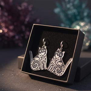 May include: A pair of cat-shaped earrings in a black gift box. The earrings are black with a white floral design. The earrings have a silver-colored hook. The gift box is black and open.