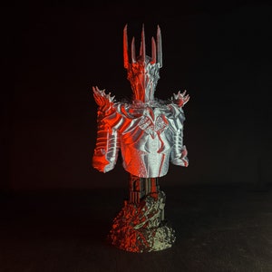 Puede incluir: Un busto plateado de una figura que lleva una corona y una armadura. La figura est&aacute; de pie sobre una base negra con textura rocosa. El busto est&aacute; iluminado con luz roja.