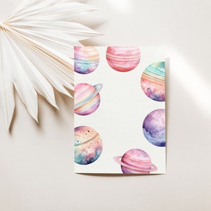 May include: Watercolor painting of planets in shades of pink, purple, blue, and green. The planets have rings and are surrounded by a white background.