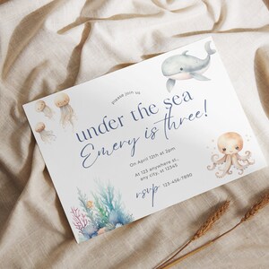 May include: A white invitation card with blue text that reads "Please join us under the sea Emery is three!"  The invitation card features a watercolor illustration of a whale, an octopus, and jellyfish. The text also includes the date, time, and location of the event.  The invitation card is on a beige fabric background.