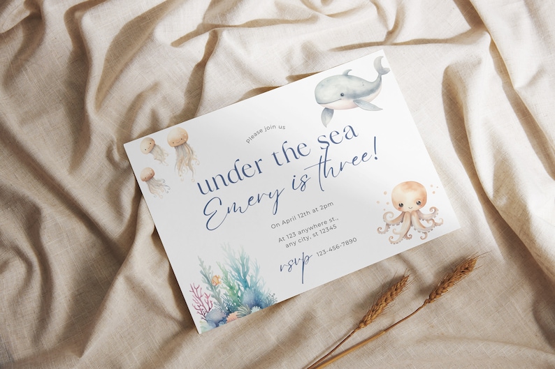 May include: A white invitation card with blue text that reads "Please join us under the sea Emery is three!"  The invitation card features a watercolor illustration of a whale, an octopus, and jellyfish. The text also includes the date, time, and location of the event.  The invitation card is on a beige fabric background.