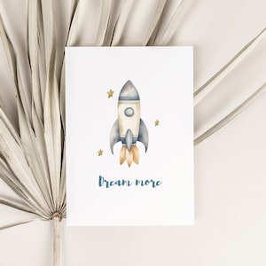 May include: A watercolor illustration of a blue and white rocket ship with orange flames, surrounded by gold stars. The text "Dream More" is written below the rocket.