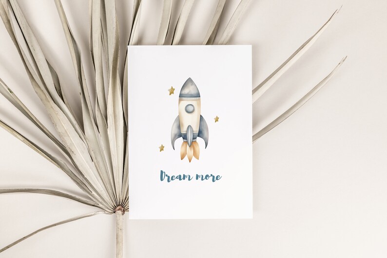 May include: A watercolor illustration of a blue and white rocket ship with orange flames, surrounded by gold stars. The text "Dream More" is written below the rocket.