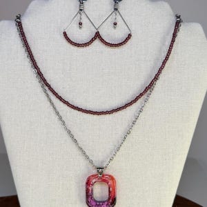May include: A layered necklace set featuring a strand of small dark red beads and a silver-tone chain with a rectangular pendant in shades of pink, orange, and purple. Matching triangular earrings feature silver-tone wire and dark red beads.