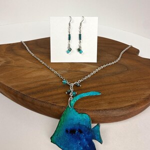 May include: A jewelry set consisting of a silver-toned chain necklace with a large, flat, fish-shaped pendant in shades of blue and green, accented with small beads. The set includes matching drop earrings featuring teal beads on silver-toned hooks.