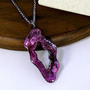 May include: A pendant necklace featuring an irregular, open-center shape with a glossy, marbled finish in shades of deep purple, magenta, and light pink. The pendant hangs from a thin, metallic chain with a subtle iridescent, oil-slick finish.