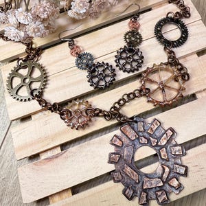 May include: A steampunk-style necklace and earring set featuring gear-shaped pendants and charms in bronze, copper, and gunmetal tones. The necklace has a chain with various gear designs, and the earrings have matching gear accents. The jewelry is displayed on a wooden surface.