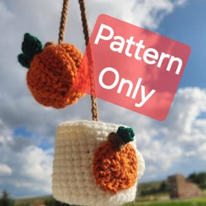 May include: Crochet pattern for a pumpkin-shaped mug cozy. The cozy is white with an orange pumpkin applique. The pattern is for sale, not the finished product. "Pattern Only" is written on a pink square.
