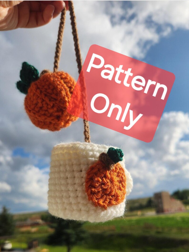 May include: Crochet pattern for a pumpkin-shaped mug cozy. The cozy is white with an orange pumpkin applique. The pattern is for sale, not the finished product. "Pattern Only" is written on a pink square.