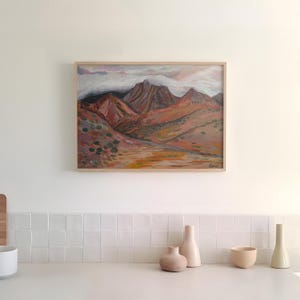 May include: A framed painting of a mountain range with clouds in the sky. The mountains are painted in shades of brown, orange, and red. The painting is hanging on a white wall above a white tiled counter with three ceramic vases and a bowl.
