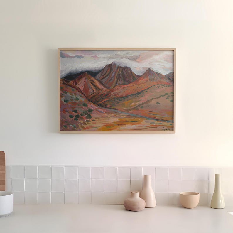 May include: A framed painting of a mountain range with clouds in the sky. The mountains are painted in shades of brown, orange, and red. The painting is hanging on a white wall above a white tiled counter with three ceramic vases and a bowl.