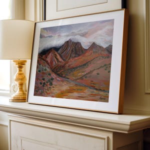 May include: A framed painting of a mountain range with a cloudy sky. The mountains are painted in shades of brown, red, and purple. The sky is painted in shades of blue, pink, and white.