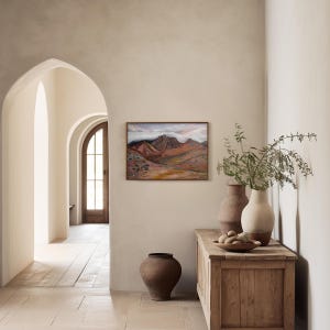 May include: A painting of a mountain range with a cloudy sky hangs on a beige wall in a room with a wooden floor. Two brown ceramic vases and a wooden cabinet with a bowl of fruit are in the foreground.