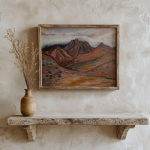 May include: A framed painting of a mountain landscape with a cloudy sky. The mountains are painted in shades of red, orange, and brown. The sky is a light blue with white clouds.